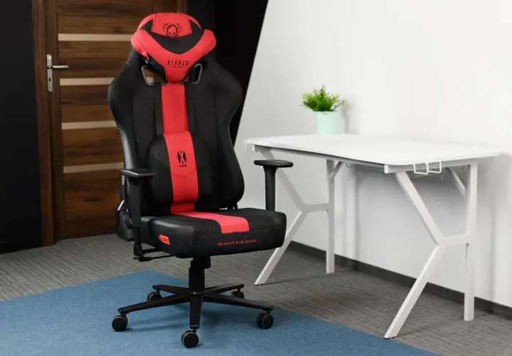 Czarno-czerwone krzesło gamingowe Diablo Chairs Player na tle białego biurka i drzwi.