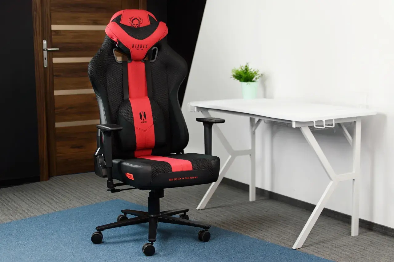 Czarno-czerwone krzesło gamingowe Diablo Chairs Player na tle białego biurka i drzwi.